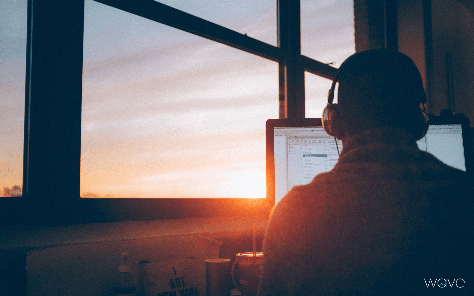 A person wearing headphones is using the computer in front of a window. The sun is setting.