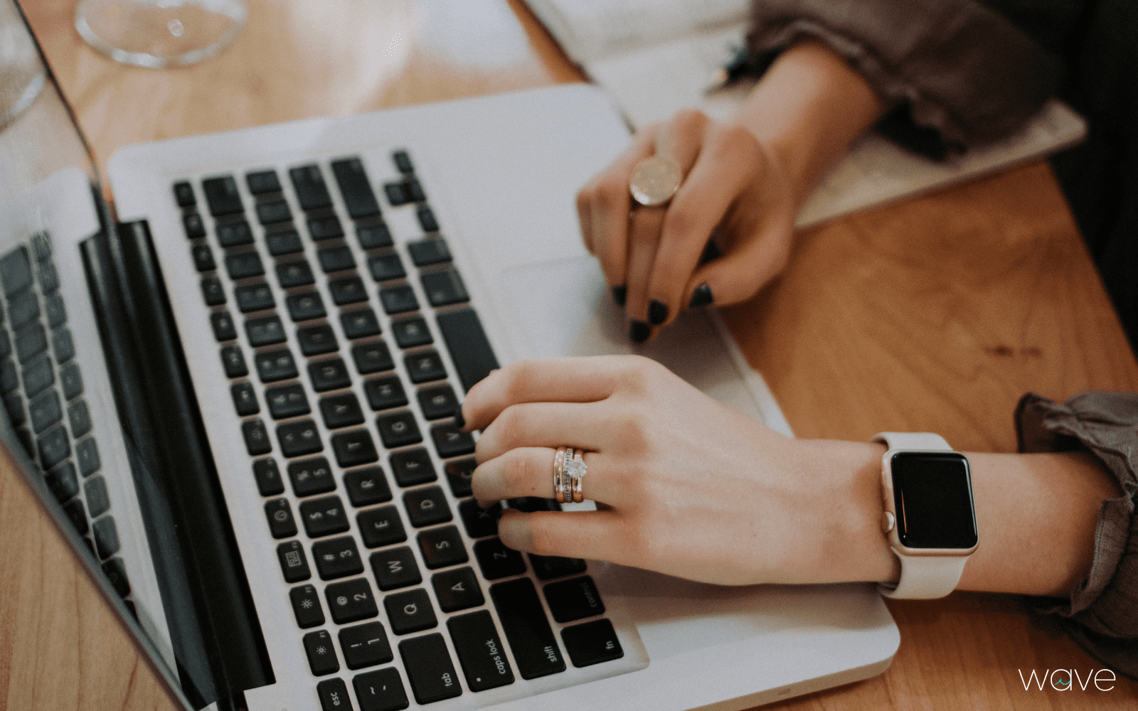 Hands wearing jewelry and watch are shown typing at a laptop.