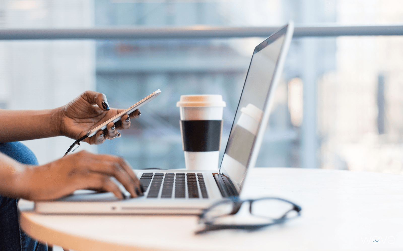 A person is sitting at a desk near a window. They are typing at a computer while also on their phone. Glasses are on the desk next to them with a cup of coffee.